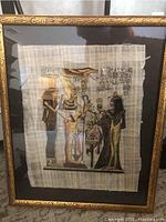 Front view of the framed Egyptian papyrus artwork showing four traditional Egyptian figures and hieroglyphs on papyrus behind glass in a carved gold-toned frame.