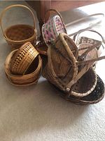 Cluster of eight assorted wicker and straw baskets of varying shapes, sizes, colors, and styles stacked together on carpeted floor.