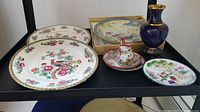 Large decorative plates, porcelain vase with stand, small creamer jug with matching plate on black shelf.