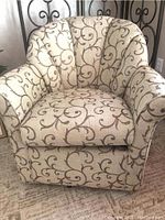 Front facing view of upholstered rotating chair with beige fabric and brown embroidered scroll pattern on fabric. Shows rounded backrest and armrests with plush seating.