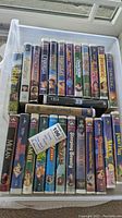 Photo of 26 Disney VHS movie tapes arranged in a container, showing titles such as Beauty and the Beast, Aladdin, Hercules, Cinderella, The Little Mermaid II, Hercules, Lady and the Tramp, The Lion King, Dumbo, and The Lion King II.