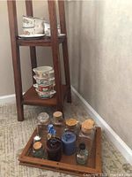 Wooden shelf holding floral gumbo bowls stacked, enamel dishes with floral pattern on top shelf, and square wooden tray on floor with several glass canisters with varying cork and screw tops.