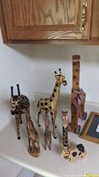 Six wooden giraffe figurines of varying sizes and styles along with one wooden cow figurine arranged on a countertop beneath a wood cabinet.