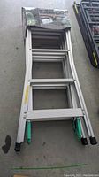 Folded aluminum multi-position ladder with green hinges and black rubber feet, partially covered with product packaging.