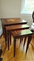 Three nested wooden nesting tables with decorative inlay tops, placed on hardwood flooring near a window providing natural light.