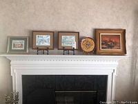 Wide view of five pieces of wall art and decorative plate arranged on a white mantle above a fireplace.