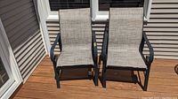Two metal frame patio chairs shown side by side on a wood deck with beige siding background. Both chairs have gray mesh fabric seating and backrest and curved metal armrests.
