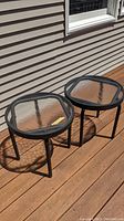 Two round patio side tables with textured glass tops and black metal frames on a wooden deck, showing light reflections on glass.