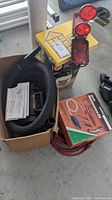 Photo shows a box containing a Metro air blower with hose and various attachments, paperwork, yellow Wolman deck sprayer box, red extension cord, Reader's Digest Do It Yourself book, and emergency roadside lights mounted on a small pole.