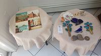 Two small tables covered in a beige cloth; one table holds four hand painted tiles with scenic views, the other table displays stained glass and decorative items.
