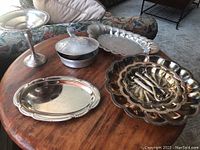 Overview of silver serving items on round wooden table showing weighted compote, unassembled tiered stand plates, silver bowls, and serving utensils.