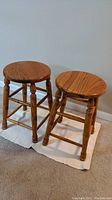 Photo showing two solid wood stools side by side with round seats and turned legs, standing on a carpeted floor with a white mat underneath.
