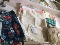 Photo showing folded assorted table linens including floral navy blue fabric, cream fringed placemats, and crochet lace pieces.