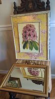 Botanical print with pink flower cluster on toile yellow background in bamboo-style white distressed wooden frame, with gilded beveled mirror below reflecting the print
