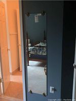 Rectangular full-length mirror hanging flush on a dark blue wall with metal corner brackets visible. Room reflection shows bed and furniture.