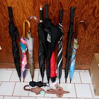 Photo showing 11 assorted umbrellas standing upright against wood panel wall, displaying mixed colors, sizes, and handle styles.
