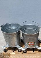 Front view of two galvanized metal oval buckets with handles placed on caster wheels, positioned on cardboard boxes. One bucket has a Behrens brand label visible.