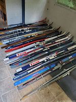 Photo showing multiple pairs of skis arranged on floor, featuring brands such as Trak, Bonna, Atomic, and others