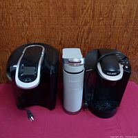 Photo showing all three Keurig coffee brewers lined up side by side on a red surface against a wood background.