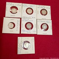 Seven Roosevelt dimes individually secured in white cardboard holders with notes on year and damage or minting defects handwritten on holders. Coins arranged on red cloth.