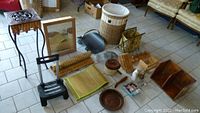 Overhead photo showing entire lot of assorted household items on tiled floor including baskets, rack, stands, framed mirror, dustpan, trays, wooden and ceramic figurines, and small furniture.