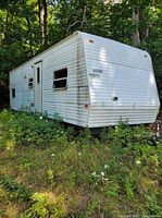 Side view of white trailer with door and windows parked in overgrown area
