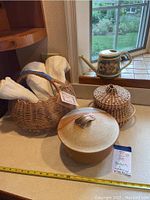 Full view of lot showing ceramic lidded bowl, bamboo basket with dish towels, smaller coiled basket, and watering can on kitchen countertop near window.