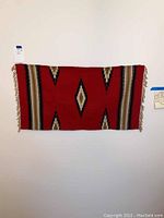 Full view of the Chimayo rug hung on a wall showing the red background with geometric diamond motifs and fringed edges.