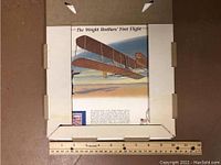 Framed print titled 'The Wright Brothers' First Flight' featuring image of early airplane in flight with ruler for scale.