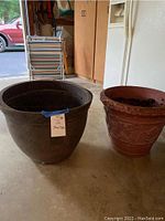 Photo showing two large planters: a black plastic one with label, and a terra-cotta one with decorative designs, placed on a garage floor near cabinets.