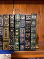 Spines of eight leather bound classic books with gilt decoration and brass shell bookend partially visible behind them