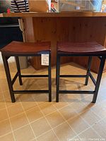 Pair of bar stools placed side by side under a counter showing black metal frames and wood toned curved seats.
