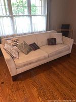Full view of sofa showing stone-colored upholstery, flared arms, dark block legs and five decorative pillows.