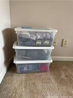 Three stacked translucent plastic bins on carpet containing yarn and knitting supplies.