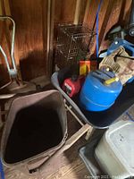 Wheelbarrow with blue propane tank, red gas can, yellow and red tin container inside, plus white plastic container and handcart visible in background inside shed