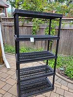 Front view of the 5-shelf black resin shelving unit standing on a tiled outdoor patio with a wooden fence background.
