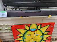 Front view of Insignia soundbar showing the fabric cover, remote control on top, and part of a Dynex TV above it, with a colorful sun artwork beneath.