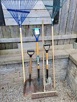 Photo showing the entire lot of three shovels, two rakes, and one push broom against a stone wall and wooden fence.