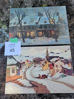 Photo showing the two laminated winter scene prints on pressed wood lying on a surface, displaying both the village street scene and the nighttime building scene.