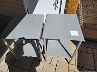 Pair of two sturdy light grey metal end tables placed outdoors on a brick patio; tables have flat tops and a lower shelf each; some discoloration visible on one top surface; no large dents or major scratches