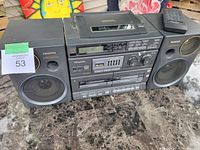 Front view of the Panasonic RX-DT680 boombox showing the central unit with tape decks, buttons, LCD display, and two detachable speakers. Remote control on top of the right speaker.