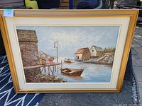 Full view of the framed oil painting showing coastal dock with boat and buildings under cloudy sky.