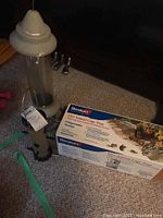 Photo shows a transparent tube bird feeder with a beige top and base next to a boxed live animal cage trap