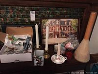 Photo showing various candlesticks including metal and ceramic holders, white taper candles, a decorative tray with a historic building image, small glass items including red goblet and clear container, and a boxed glass owl figurine.
