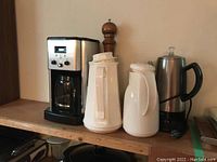 Photo showing the four items: electric coffee pot, two white ceramic thermos carafes, and wooden pepper grinder on a wooden shelf.