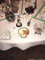 Table with porcelain and glass figurines, decorative plate, glass bowl, and metal box arranged on a table with a white padded cloth.