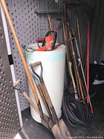 Photo of garden tools showing two shovels with wooden handles leaning against a white cylindrical container, with a large orange hedge trimmer on top and a coiled black hose nearby.