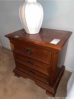 Front angled view of the cherry wood nightstand showing three drawers with metal handles and a large white ceramic vase on top.