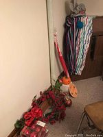 Photo showing a red Christmas wreath with red berries and a large red bow resting against a wall with some Halloween decorations nearby, including a pumpkin bucket and round orange decoration, plus a hanging colorful striped cloth item (clown costume) and folded beach umbrella in cover.