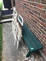 Three items placed along a brick wall: two folded vintage lawn chairs with white metal frames and fabric slats in shades of brown and cream, and one green wooden bench with weathered paint.